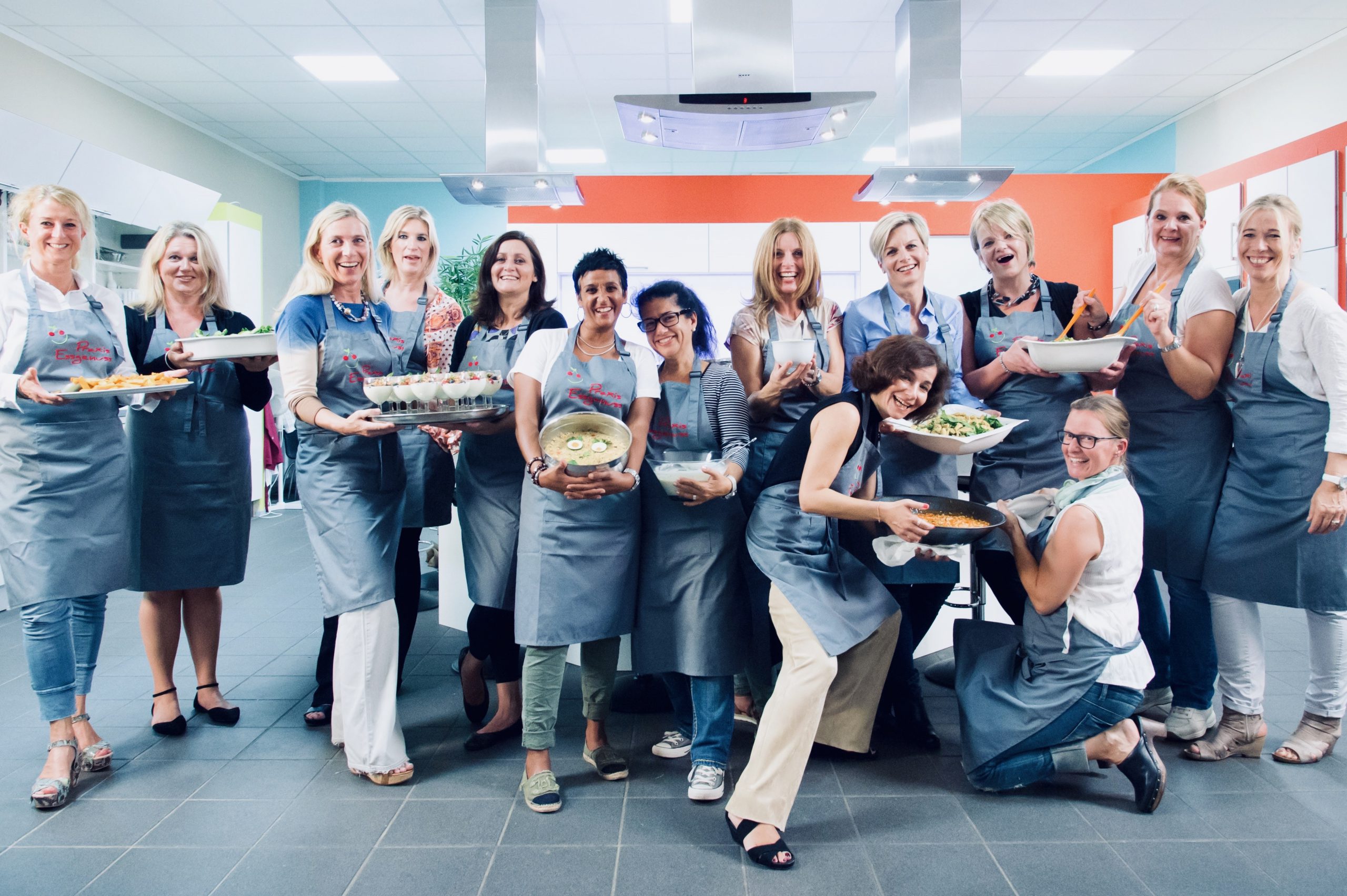 Eine Gruppe von Frauen in Praxis Essgenuss Schürze in den Hoster Küchenstudios in Krefeld. Die Frauen halten unterschiedliche, selbst zubereitete Lebensmittel in den Händen und lachen.